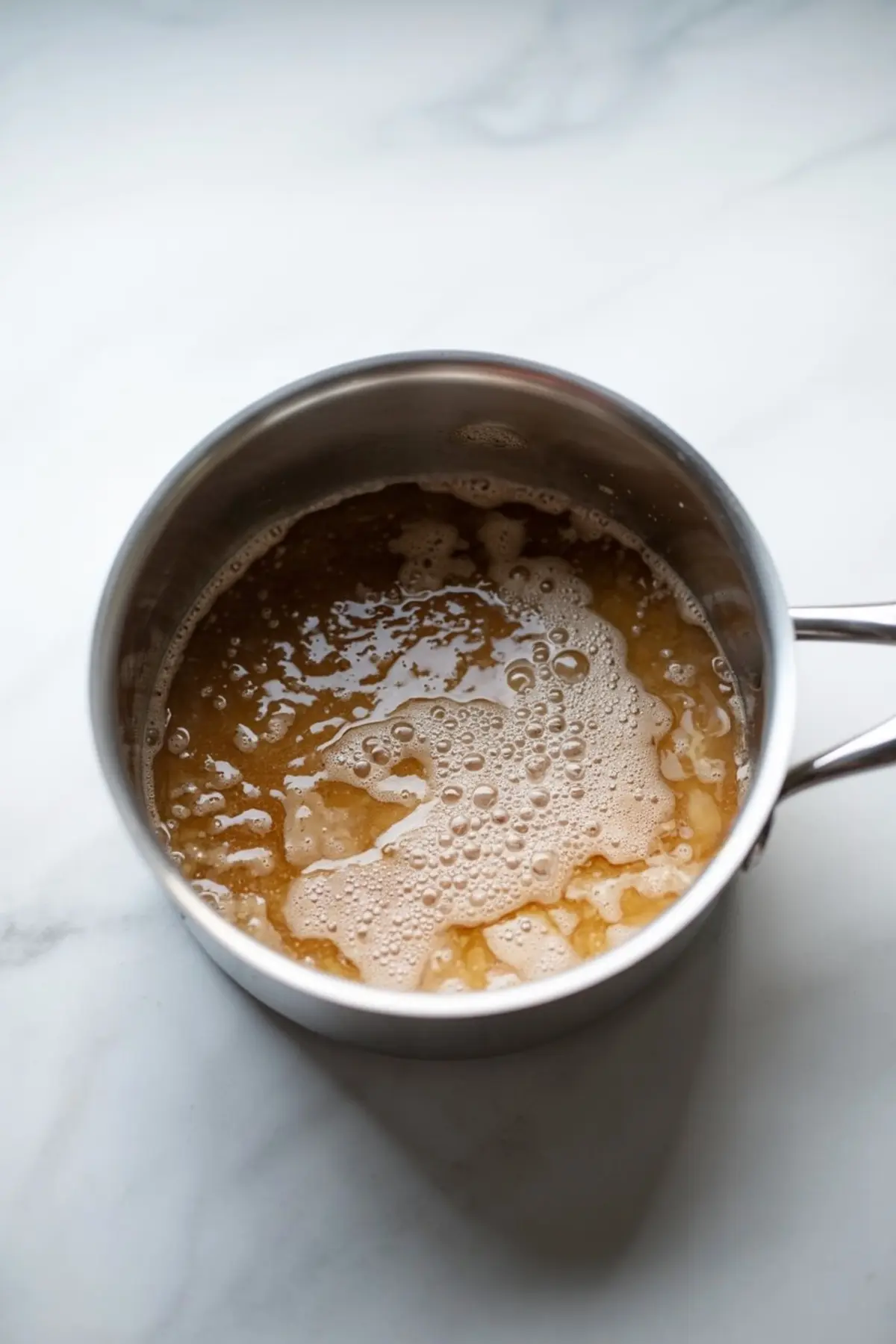 Apple pie filling mixture simmering in a stainless steel saucepan, showing a bubbling golden-brown texture as it cooks on a marble counter.