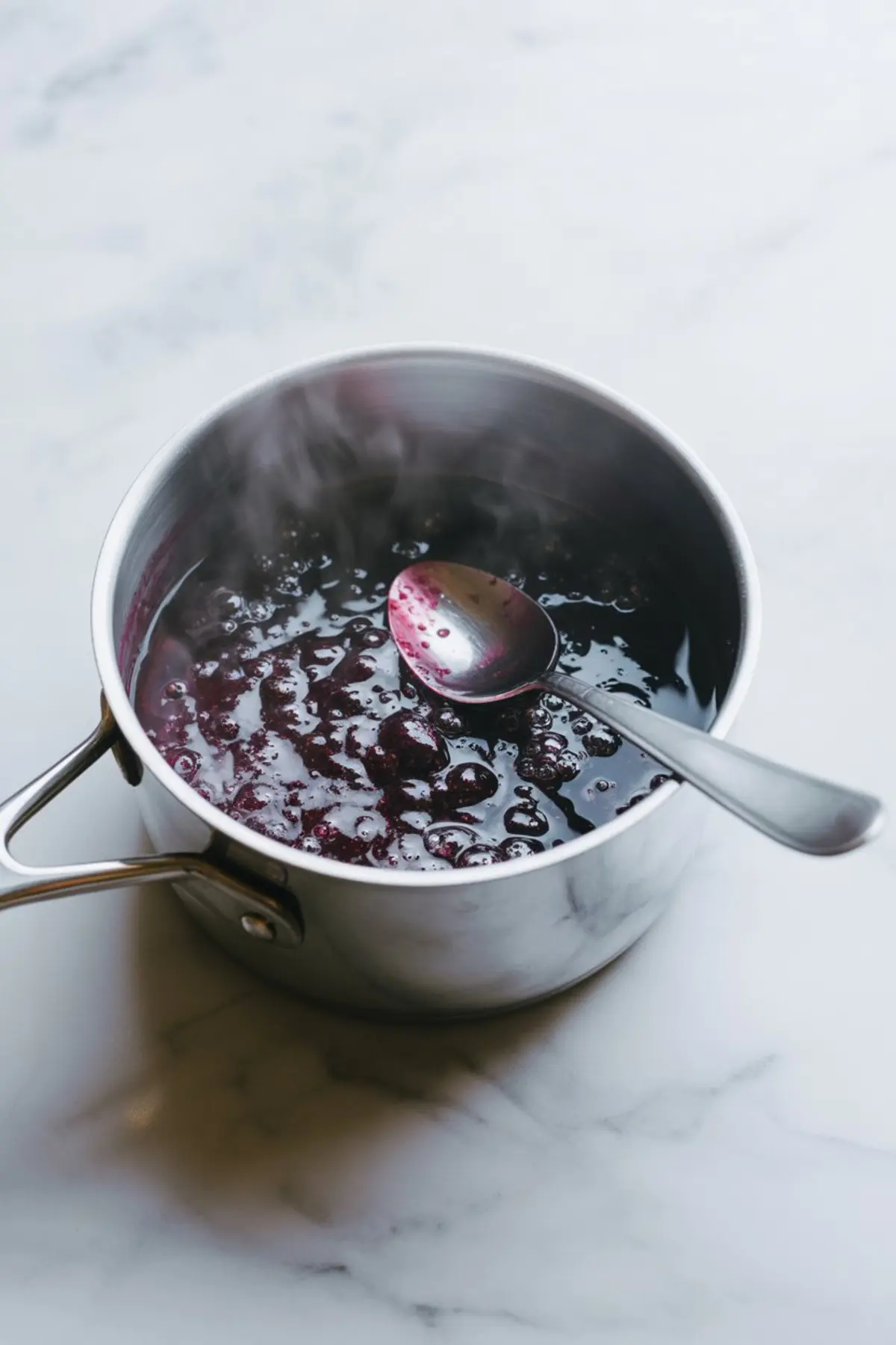Hot bubbling blackberry jam cooking in a stainless steel pot with steam rising, showing a metal spoon resting inside on a marble surface.