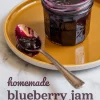 Glass jar of thick blueberry jam on a golden plate with a vintage silver spoon coated in jam placed beside it, set on a marble background with overlay text reading “homemade blueberry jam recipe.”