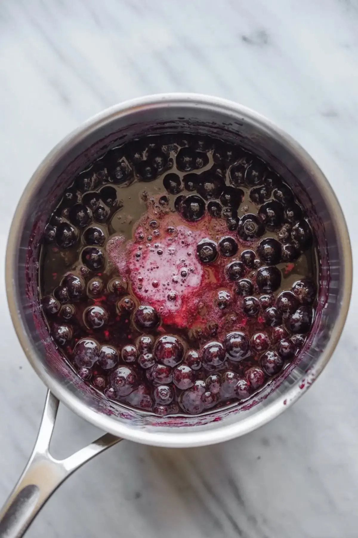 Overhead view of a stainless steel saucepan filled with bubbling blueberry sauce in the process of cooking, showing plump whole berries and deep purple juices on a marble surface.