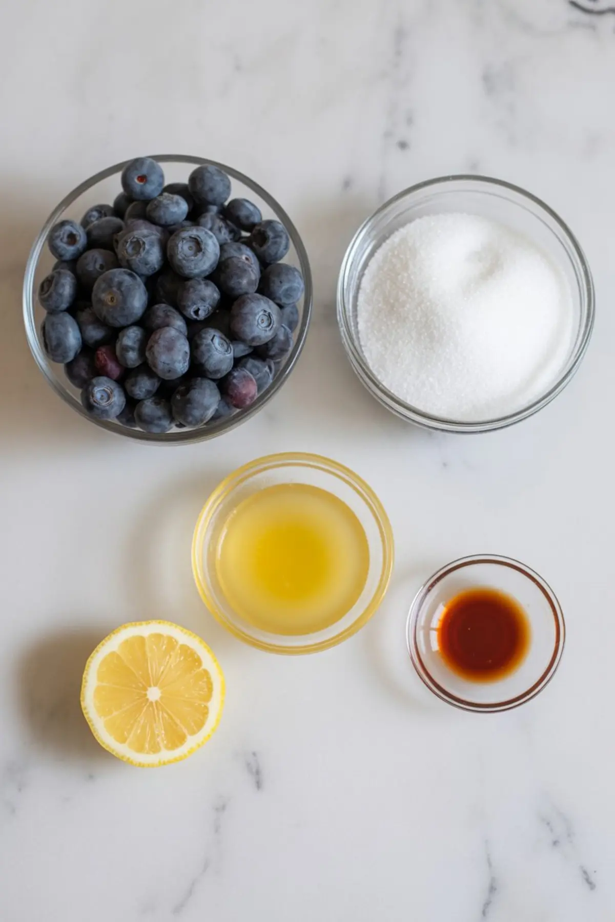 Flat lay of fresh blueberry sauce ingredients on a white countertop, including a glass bowl of blueberries, a bowl of white granulated sugar, lemon juice, vanilla extract, and a halved lemon.