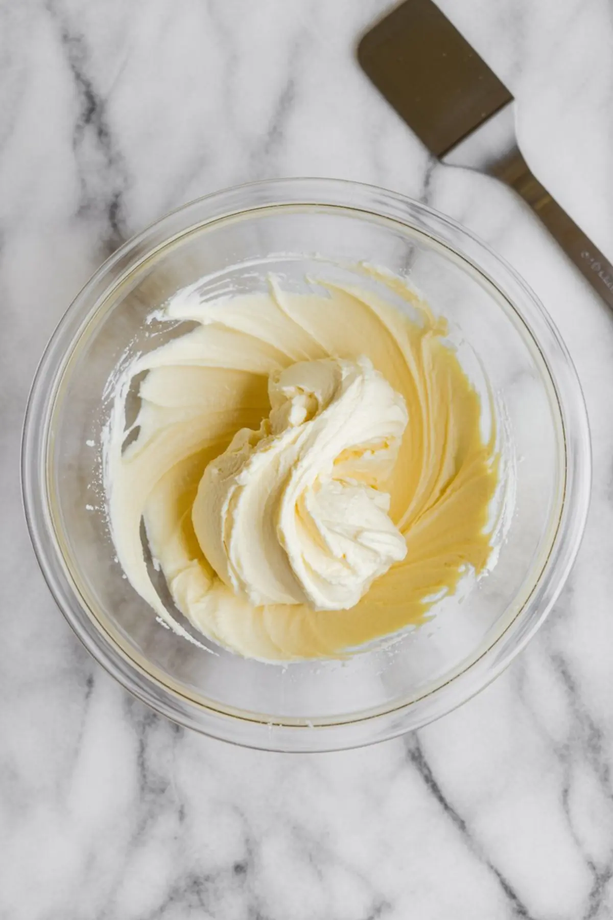 Whipped cream cheese mixture in a glass mixing bowl on a marble surface with a spatula beside it.
