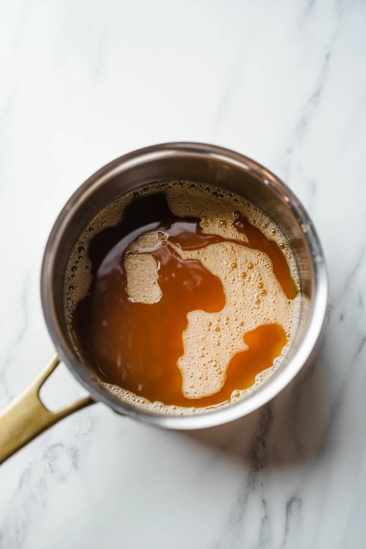 Saucepan filled with golden brown caramel bubbling on the surface, sitting on a white marble counter, illustrating the cooking process for homemade caramel sauce.