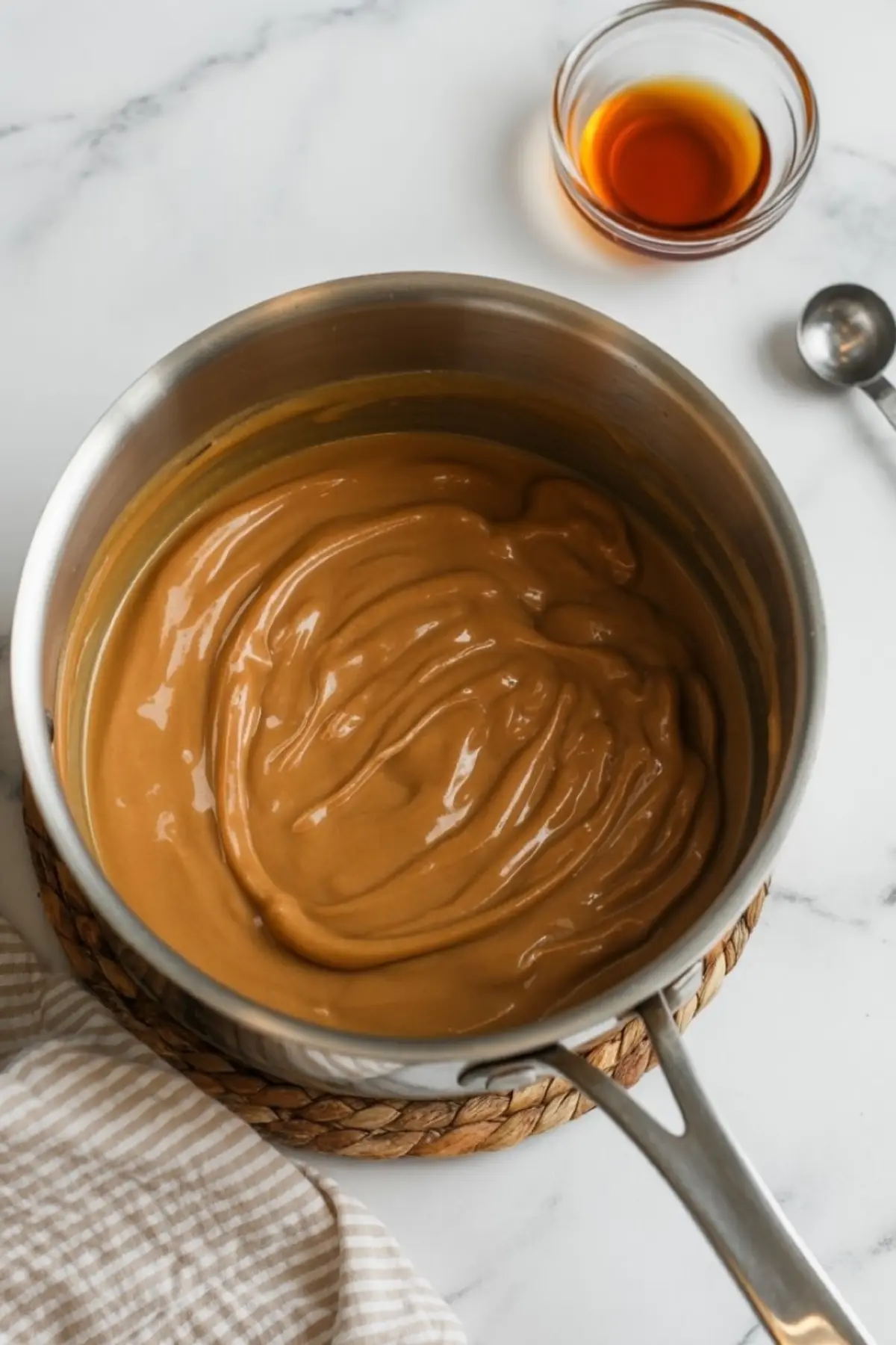 A saucepan filled with thick, golden-brown caramel sauce sits on a woven trivet over a marble surface, with a small glass bowl of vanilla extract and a metal measuring spoon nearby.