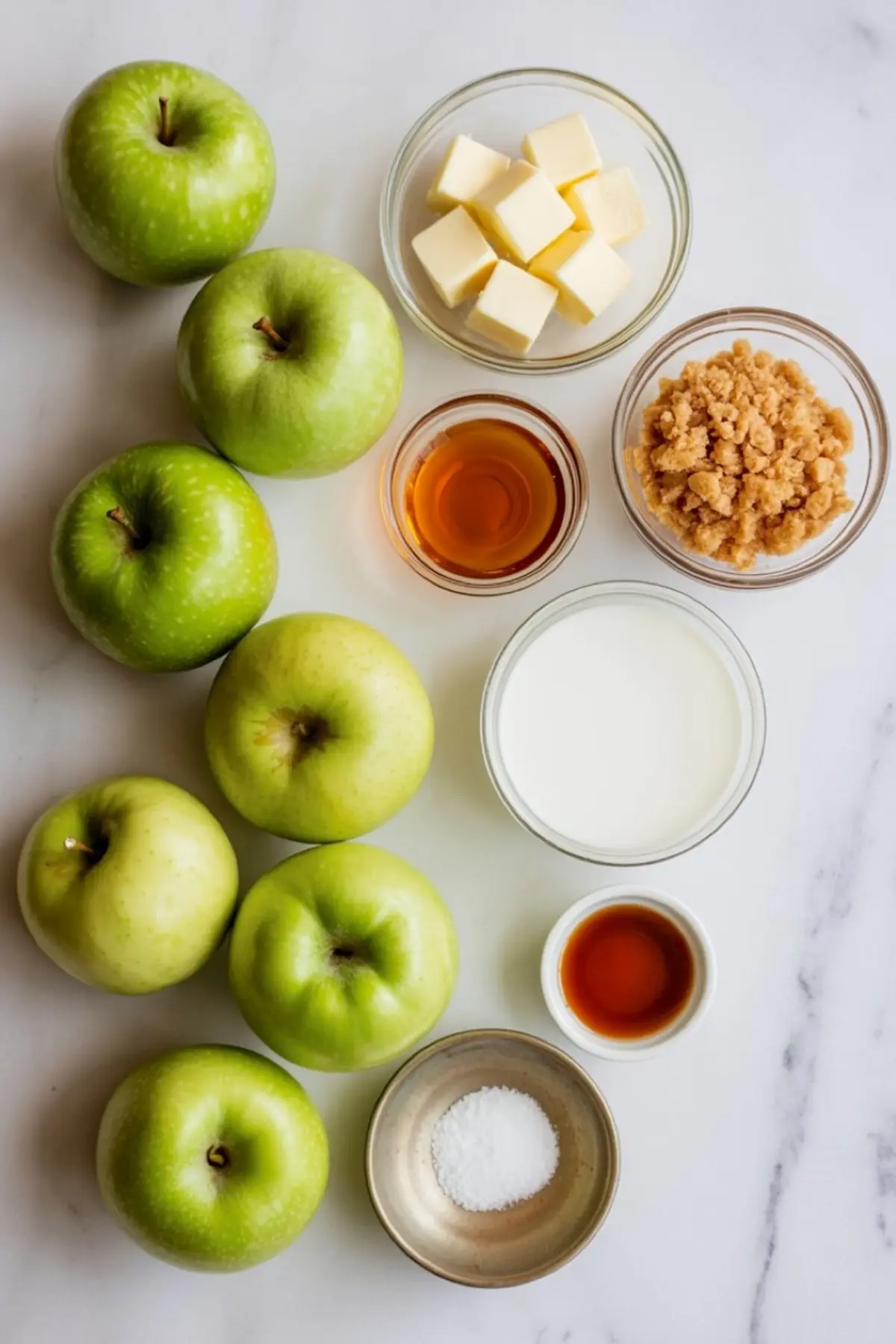 A flat lay of ingredients for homemade caramel apples, including fresh green apples, cubed butter, brown sugar, heavy cream, vanilla extract, salt, and corn syrup arranged on a white marble countertop.