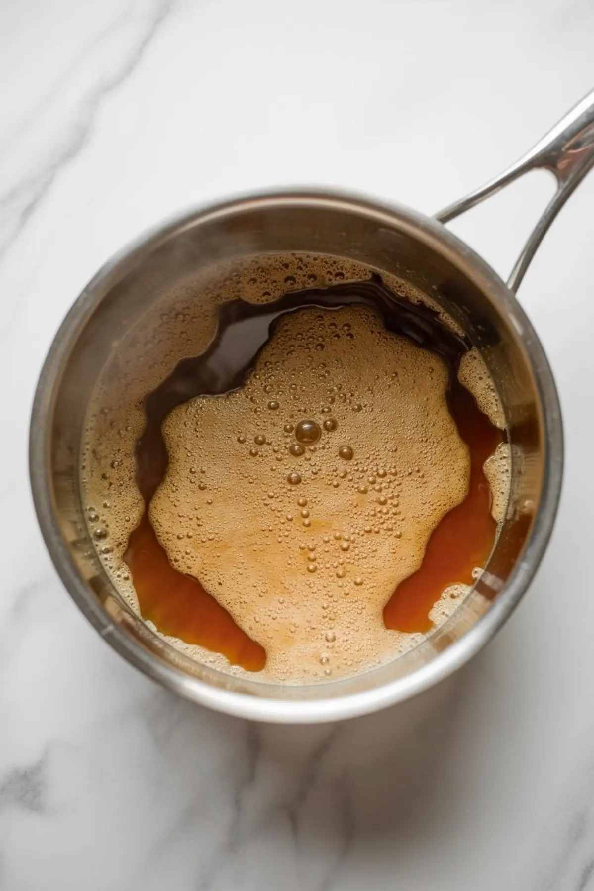 A top-down view of bubbling caramel cooking in a stainless steel saucepan, with amber tones and visible foam on the surface indicating the caramelization process.