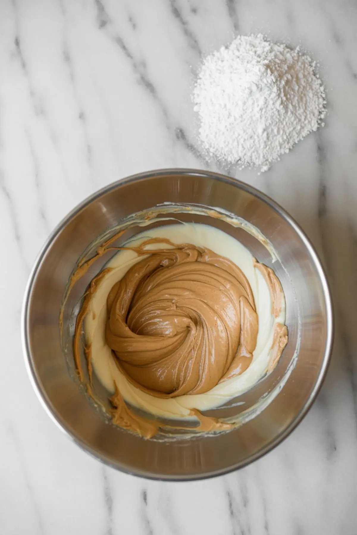 Swirled peanut butter and cream cheese mixture in a metal bowl with a mound of powdered sugar beside it on a marble counter.