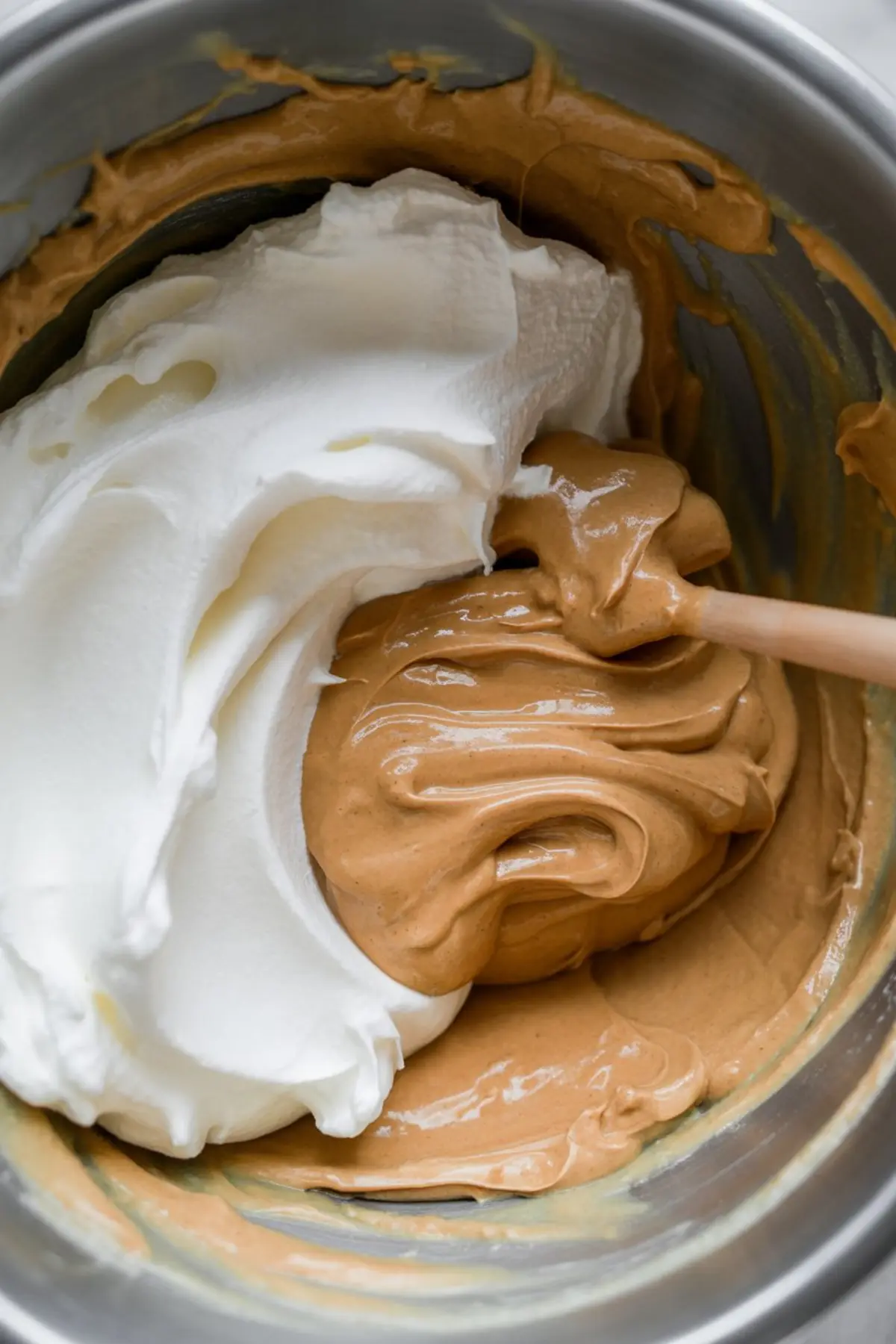 Whipped cream and peanut butter mixture being folded together in a stainless steel mixing bowl with a spatula for a creamy dessert filling.