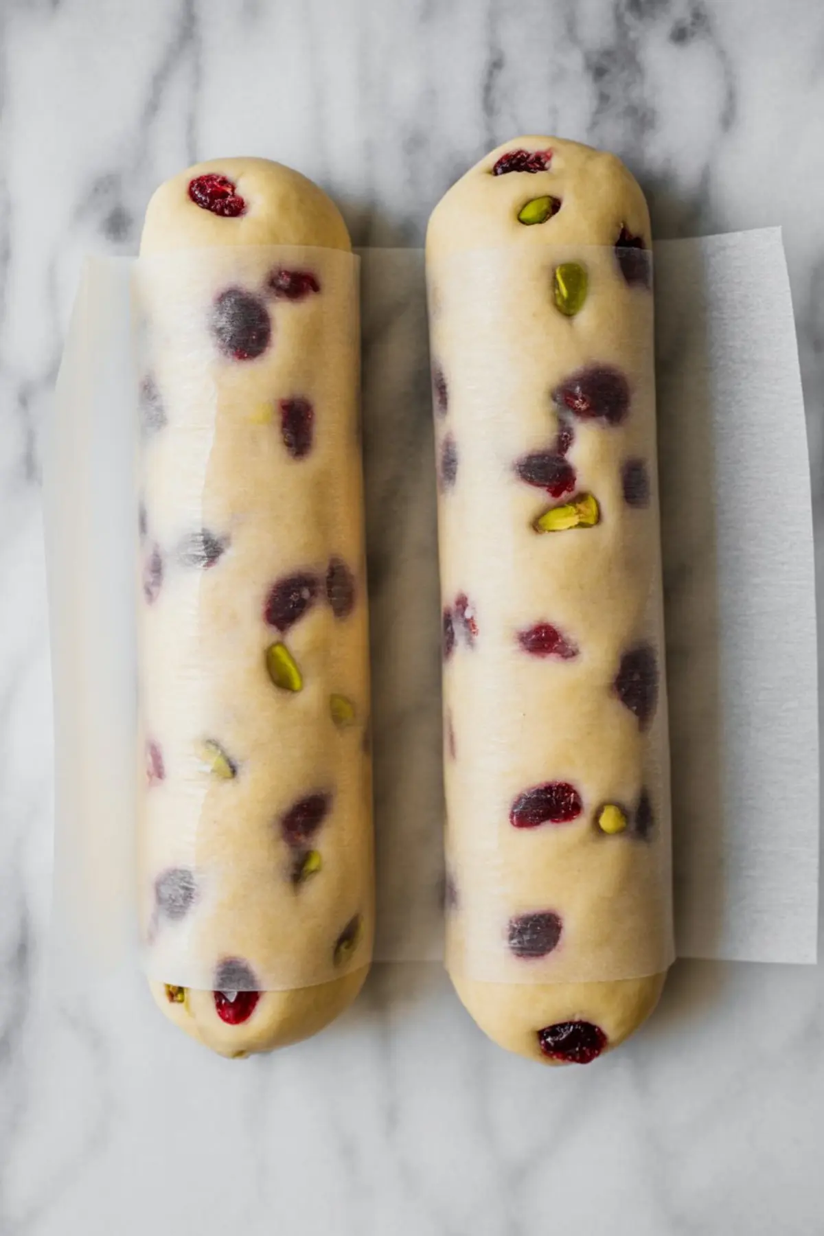 Two cylindrical logs of shortbread dough filled with chopped pistachios and dried cranberries are wrapped in parchment paper, prepped for chilling and slicing.
