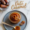 Flat lay of a small glass jar filled with thick, swirled date caramel on a gray plate, accompanied by a vintage spoon, a halved date, and a bowl of whole Medjool dates in the background. Text overlay reads “3 ingredient Date Caramel.”