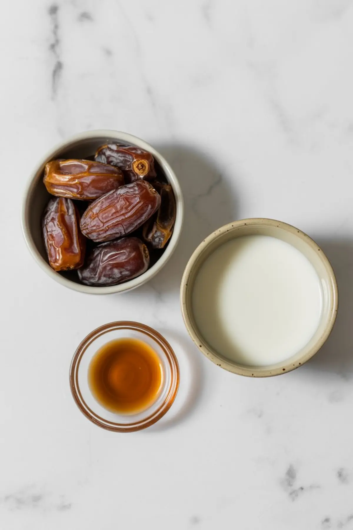 Overhead view of three small bowls on a marble surface containing the ingredients for date caramel: whole Medjool dates, creamy plant-based milk, and amber vanilla extract.