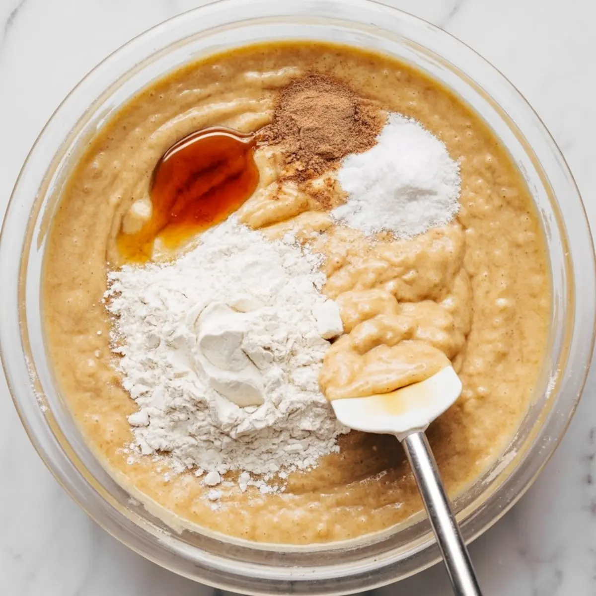 Mixing bowl with thick batter, flour, vanilla extract, baking soda, and cinnamon being folded in with a white spatula for dessert recipe preparation.