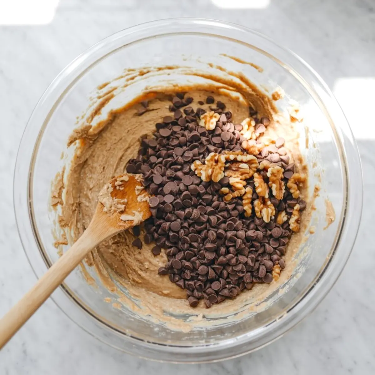 Mixing bowl filled with cookie dough or pie batter, topped with chocolate chips and chopped walnuts being stirred with a wooden spoon in natural light.