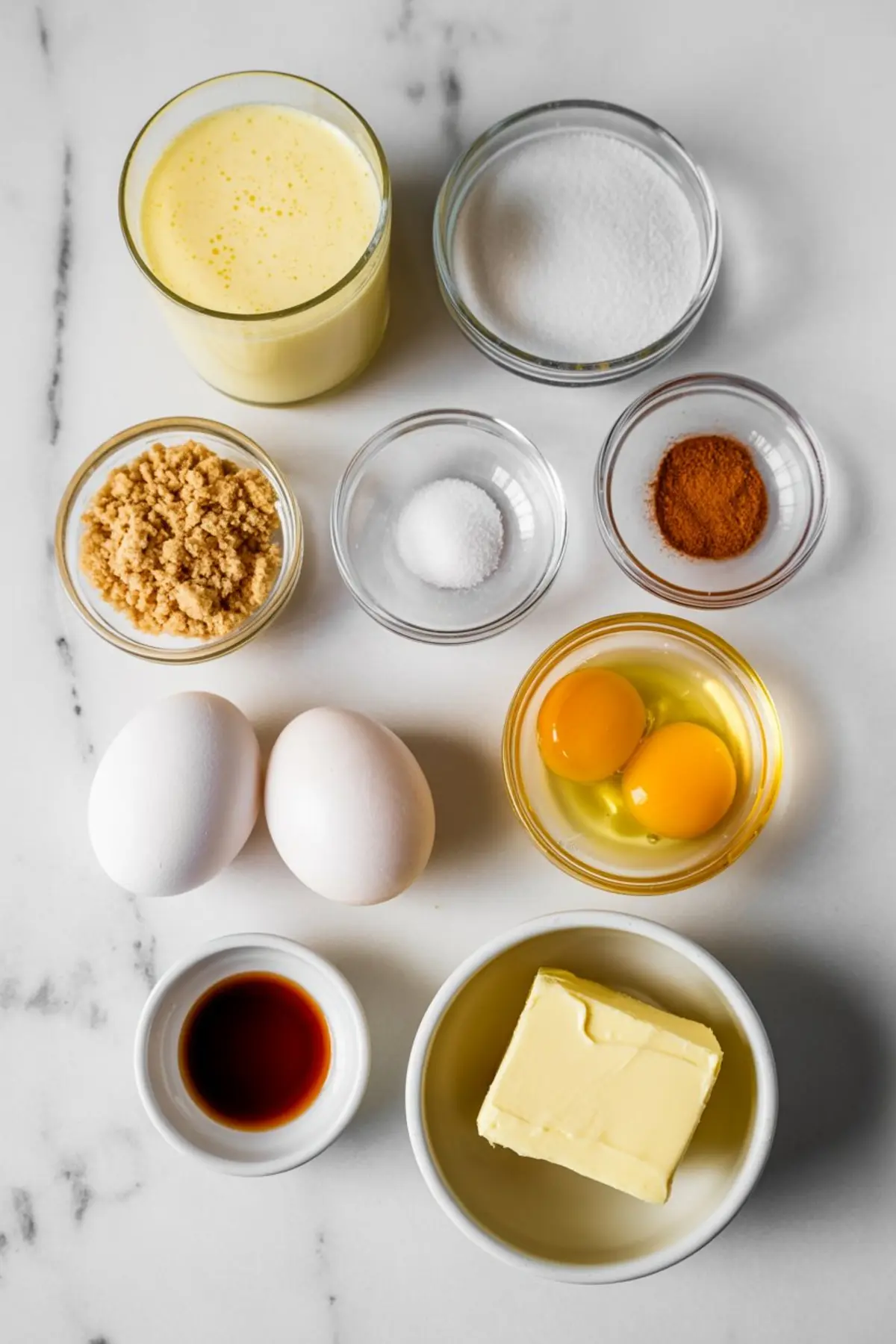 Overhead view of ingredients for eggnog pie including eggs, butter, sugar, brown sugar, nutmeg, salt, vanilla extract, and eggnog, all arranged in small glass and ceramic bowls.
