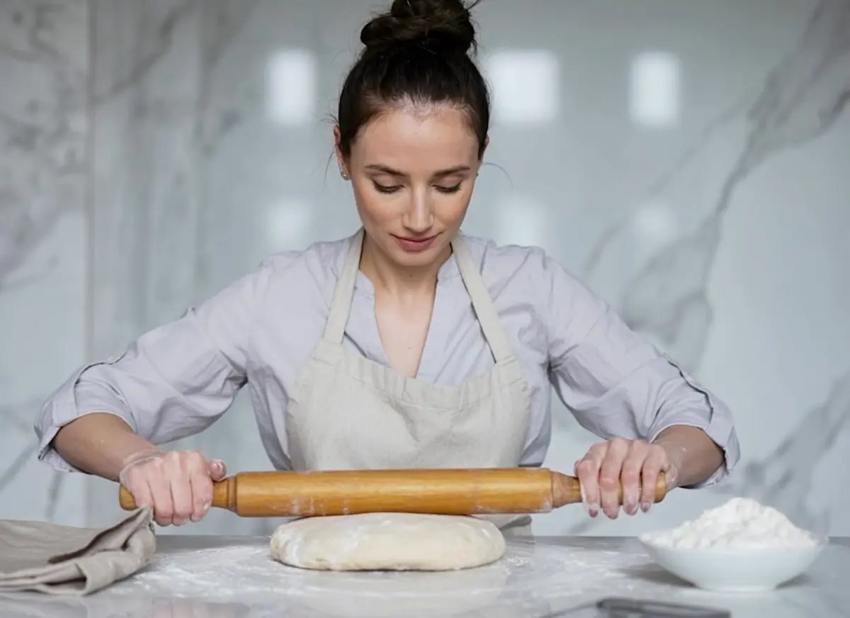 Emma wearing a light gray apron rolls out dough with a wooden rolling pin on a floured countertop ina Emma's cake studio, with a bowl of flour beside her.