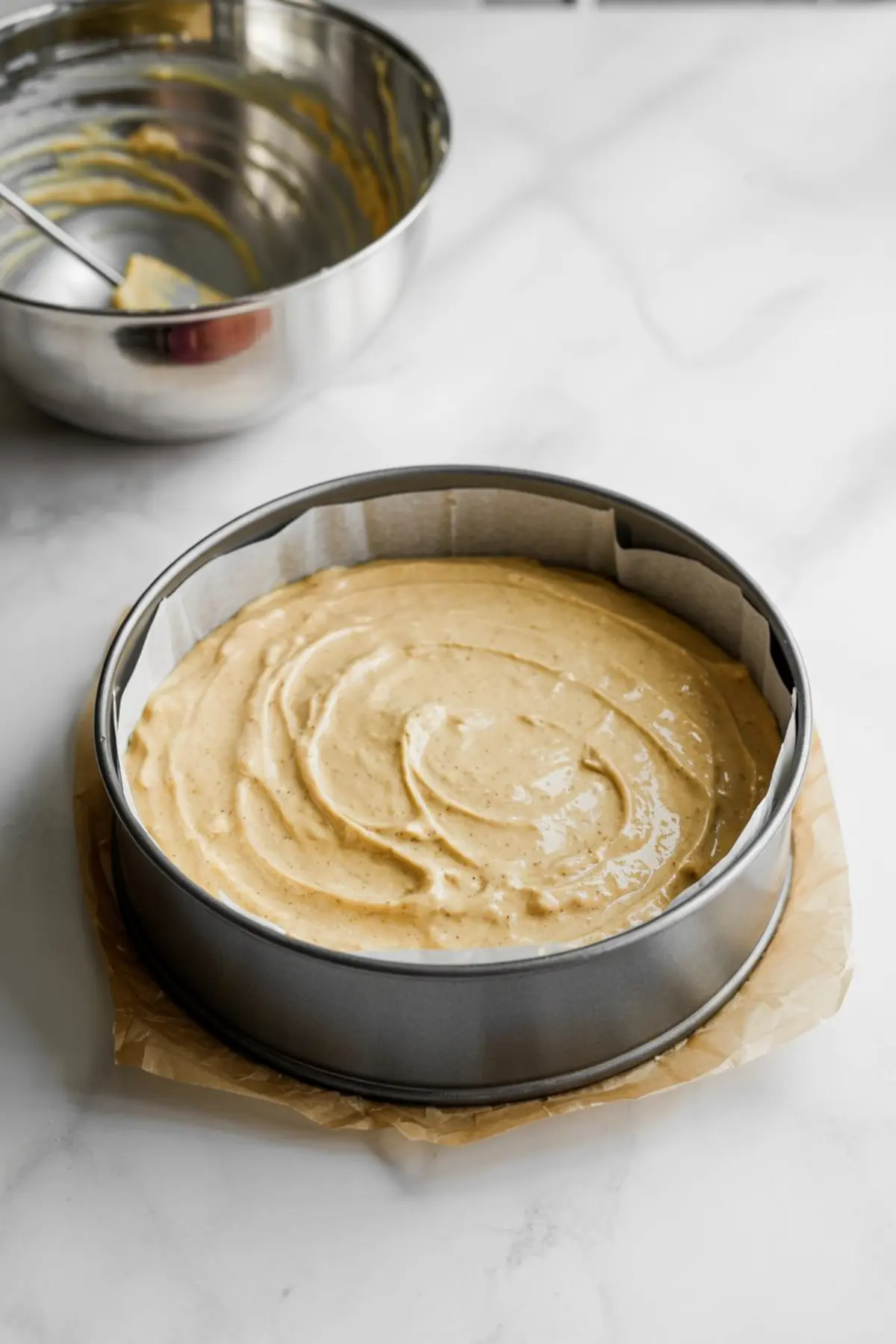 A round cake pan lined with parchment paper is filled with smooth fig cake batter, ready for baking, with an empty mixing bowl in the background.