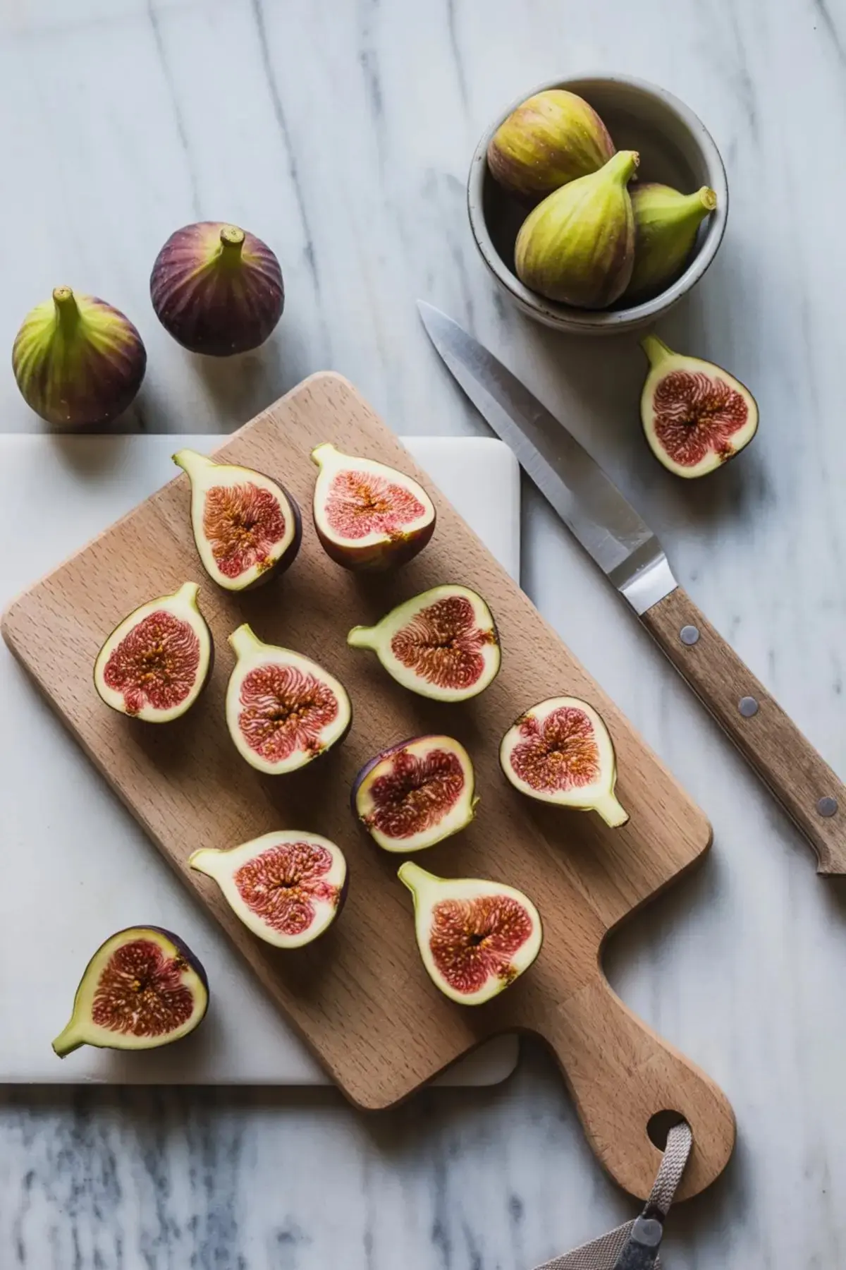 Fresh figs halved on a wooden cutting board beside a knife and small bowl, highlighting their rich color and seedy interior on a marble background.