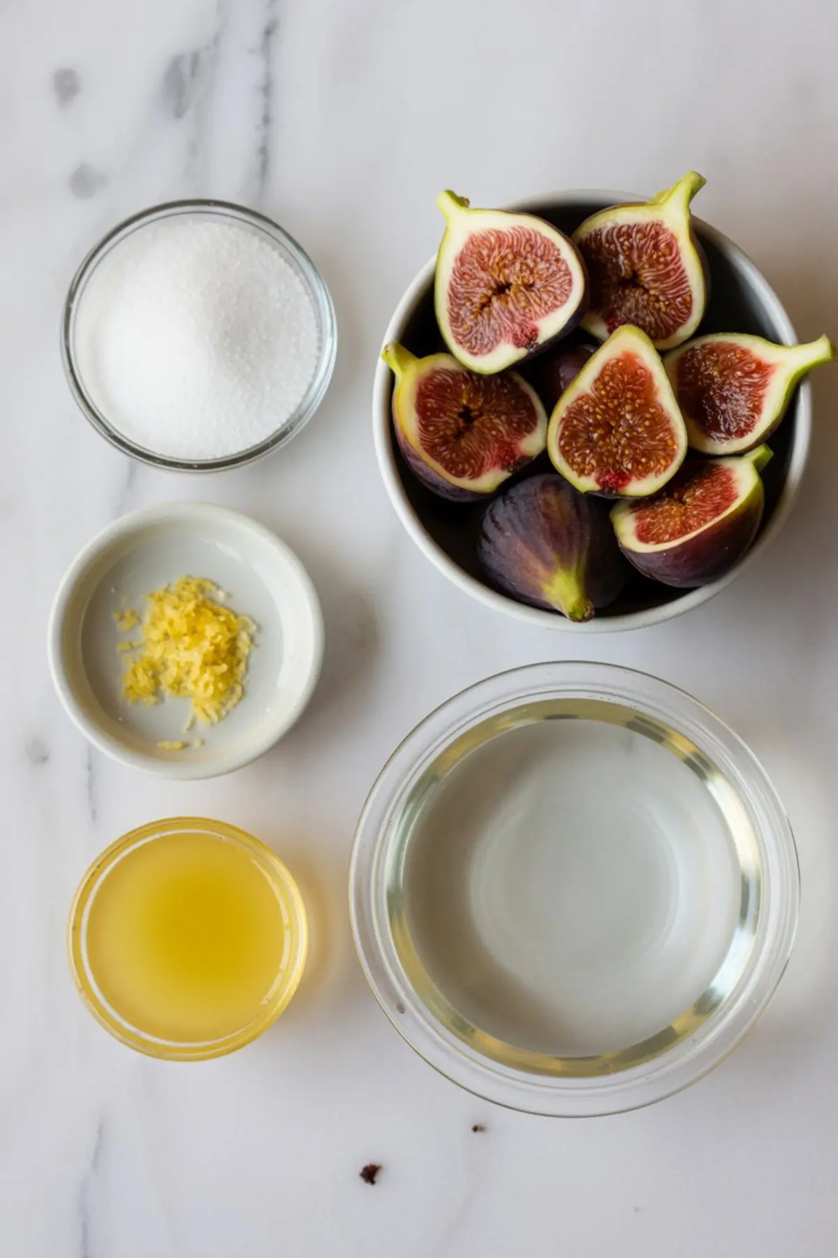 Flat lay of fig preserves ingredients including fresh halved figs, granulated sugar, lemon juice, lemon zest, and white vinegar on a white marble surface.