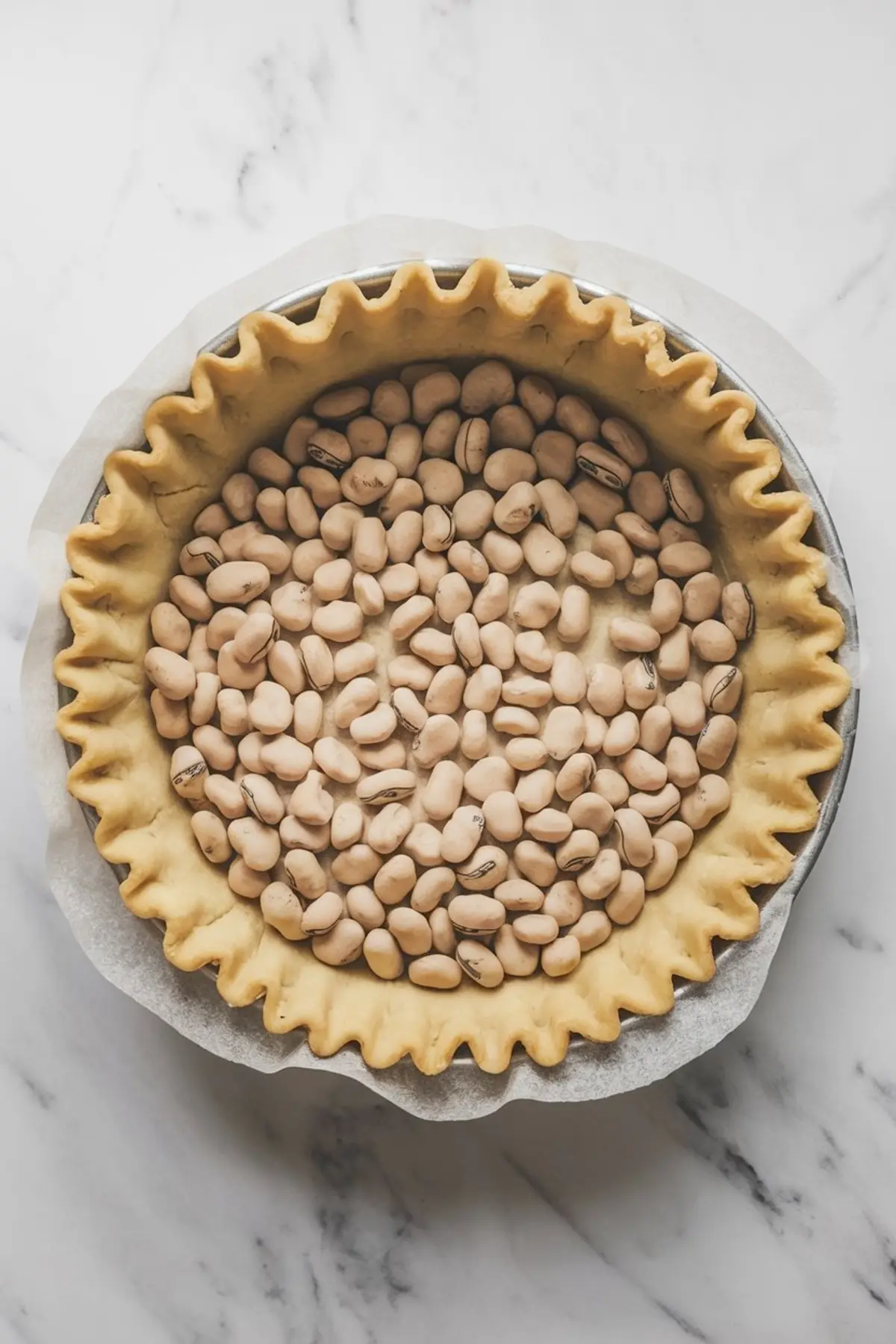 Pie crust in a metal tin lined with parchment paper and filled with dried beans for blind baking, showing a close-up of the crimped dough edges.