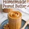 A glass jar filled with smooth homemade peanut butter on a blue plate, surrounded by shelled peanuts and a wooden spoon, with a text overlay reading “How to make Homemade Peanut Butter.”