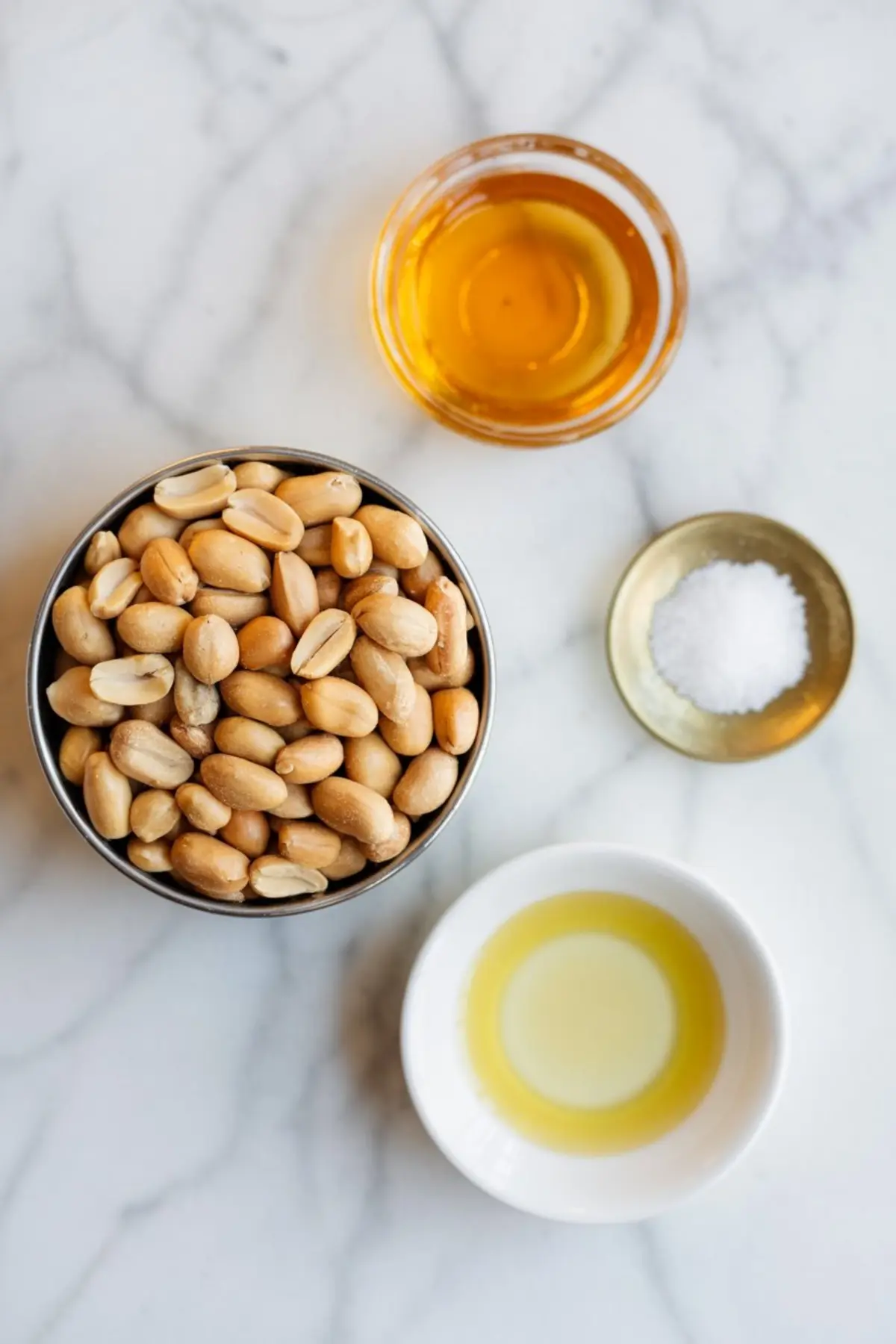 Overhead view of peanut butter ingredients on a marble surface including raw shelled peanuts in a metal bowl, honey in a glass dish, salt in a brass dish, and oil in a white ceramic bowl.