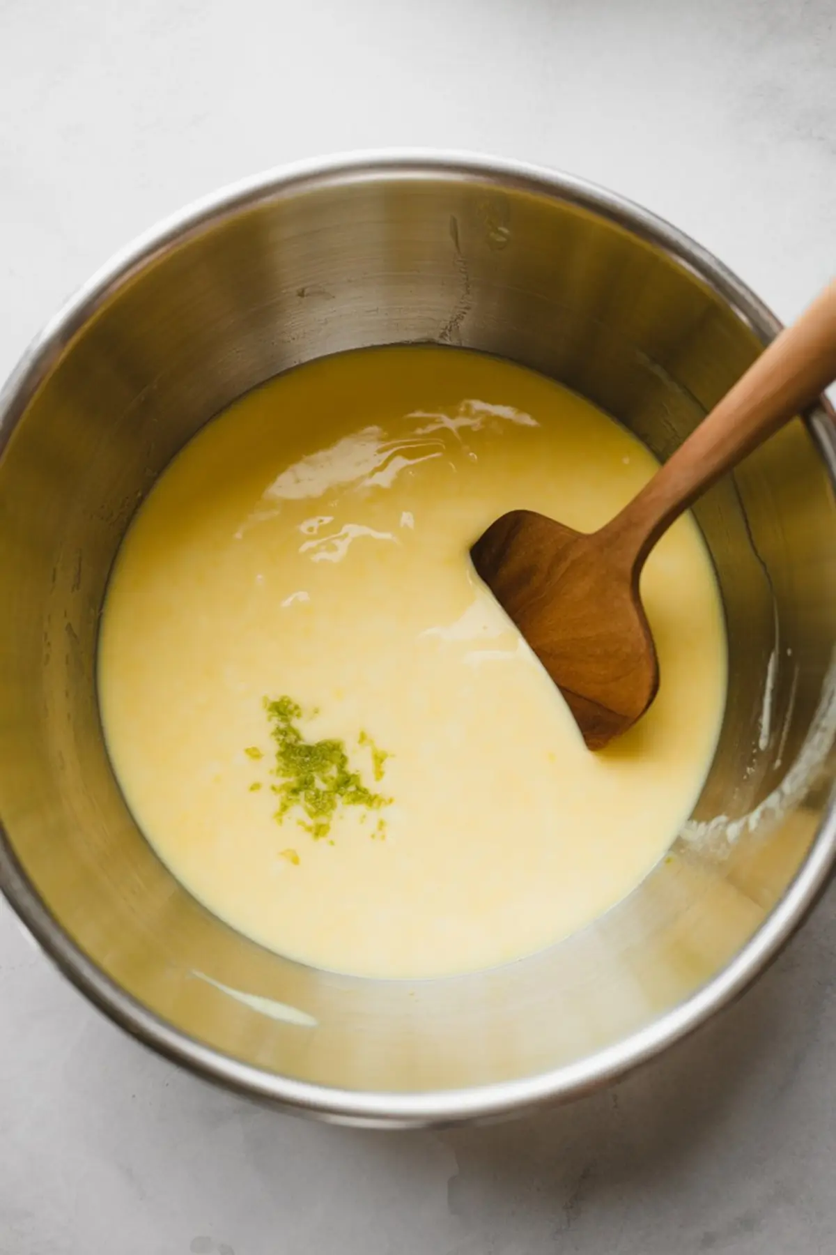 Stainless steel mixing bowl filled with creamy yellow key lime filling, topped with fresh lime zest and a wooden spatula, capturing the citrus dessert-making process.