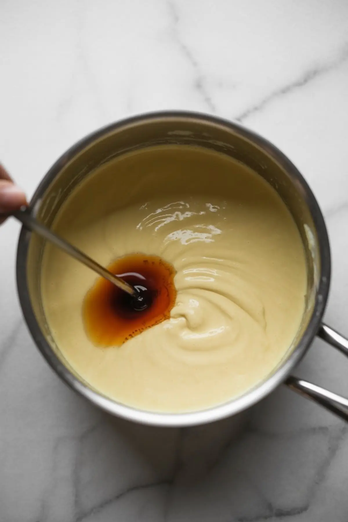 Saucepan filled with smooth panna cotta cream being mixed with a swirl of vanilla extract on a white marble surface.