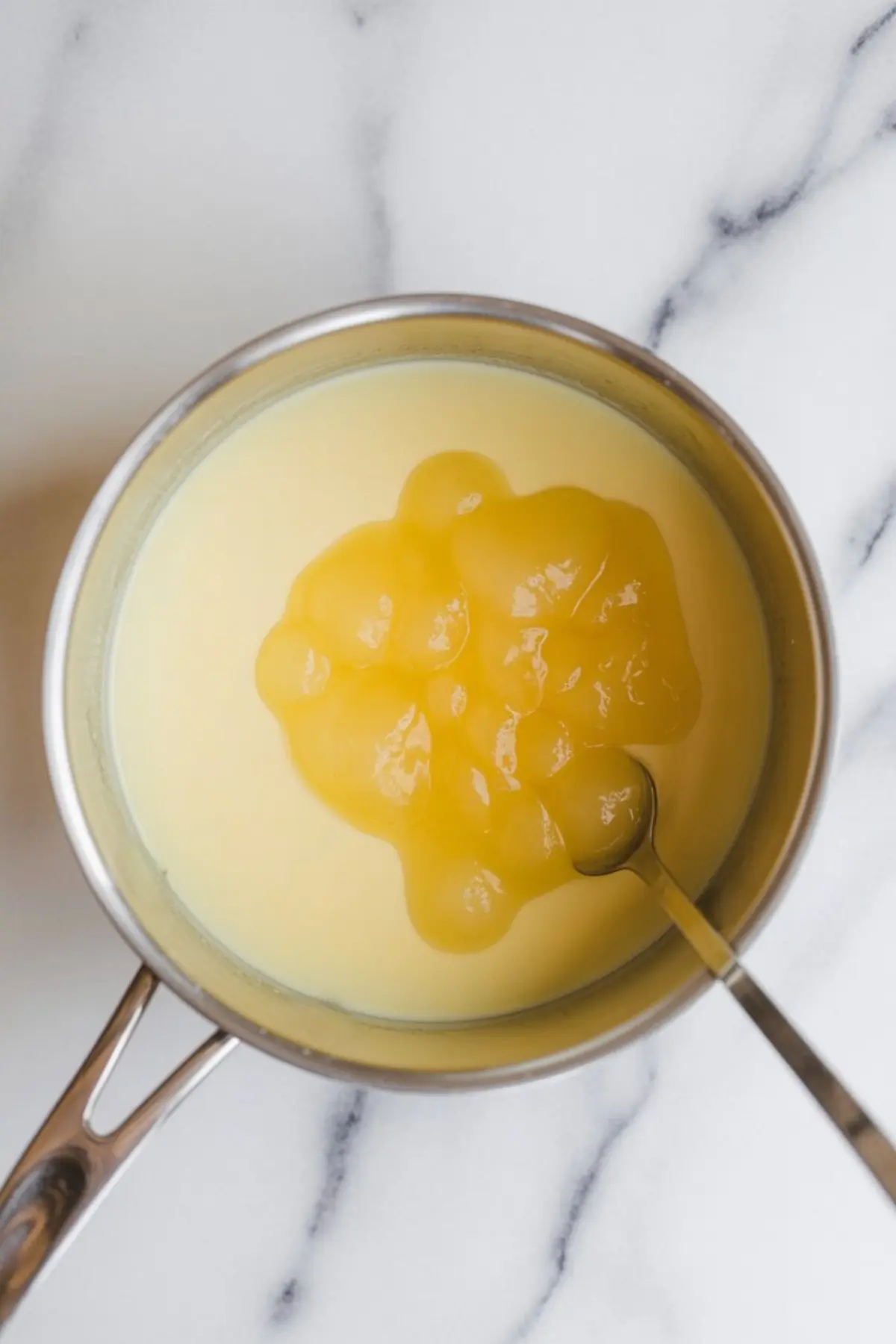 Saucepan of warm panna cotta mixture with a spoon stirring in lemon curd, creating a rich and creamy citrus base.