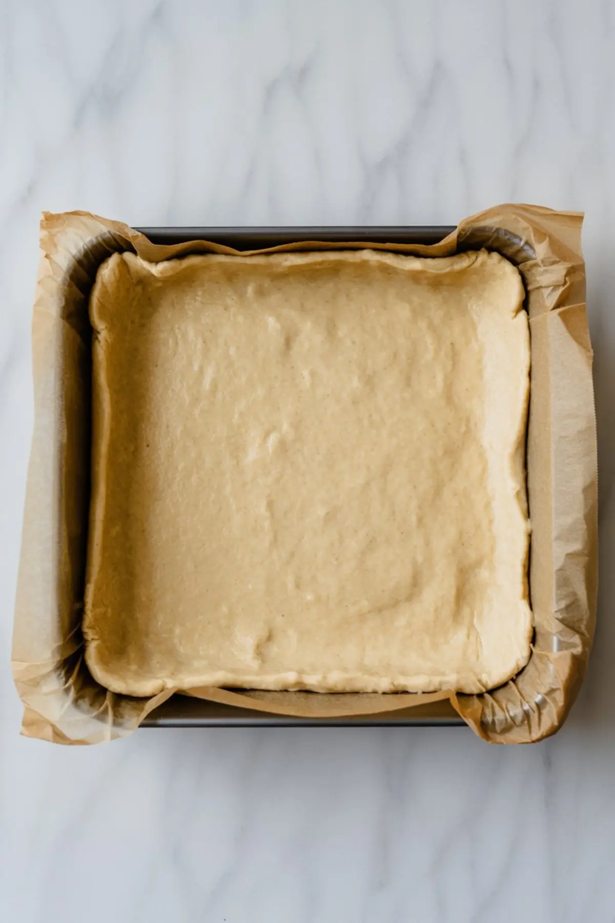 Unbaked shortbread crust pressed evenly into a square parchment-lined baking pan, ready to be used as the base layer for apple pie bars.