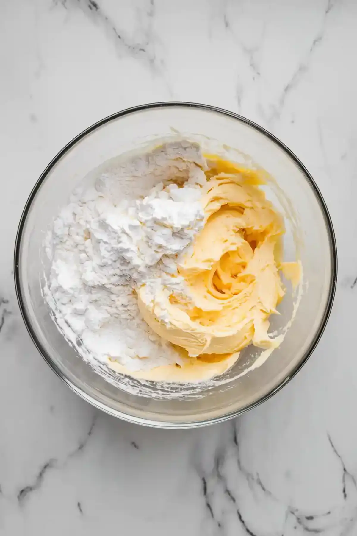 Glass bowl of whipped butter and powdered sugar mixture on a marble counter, early stage of cookie dough preparation.
