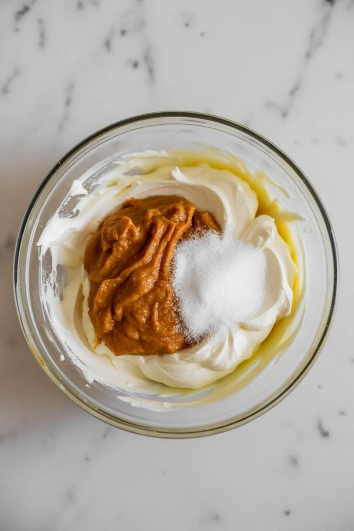 Overhead view of a mixing bowl with cream cheese, pumpkin purée, and granulated sugar on a white marble surface, ready to be blended for a fall dessert recipe.