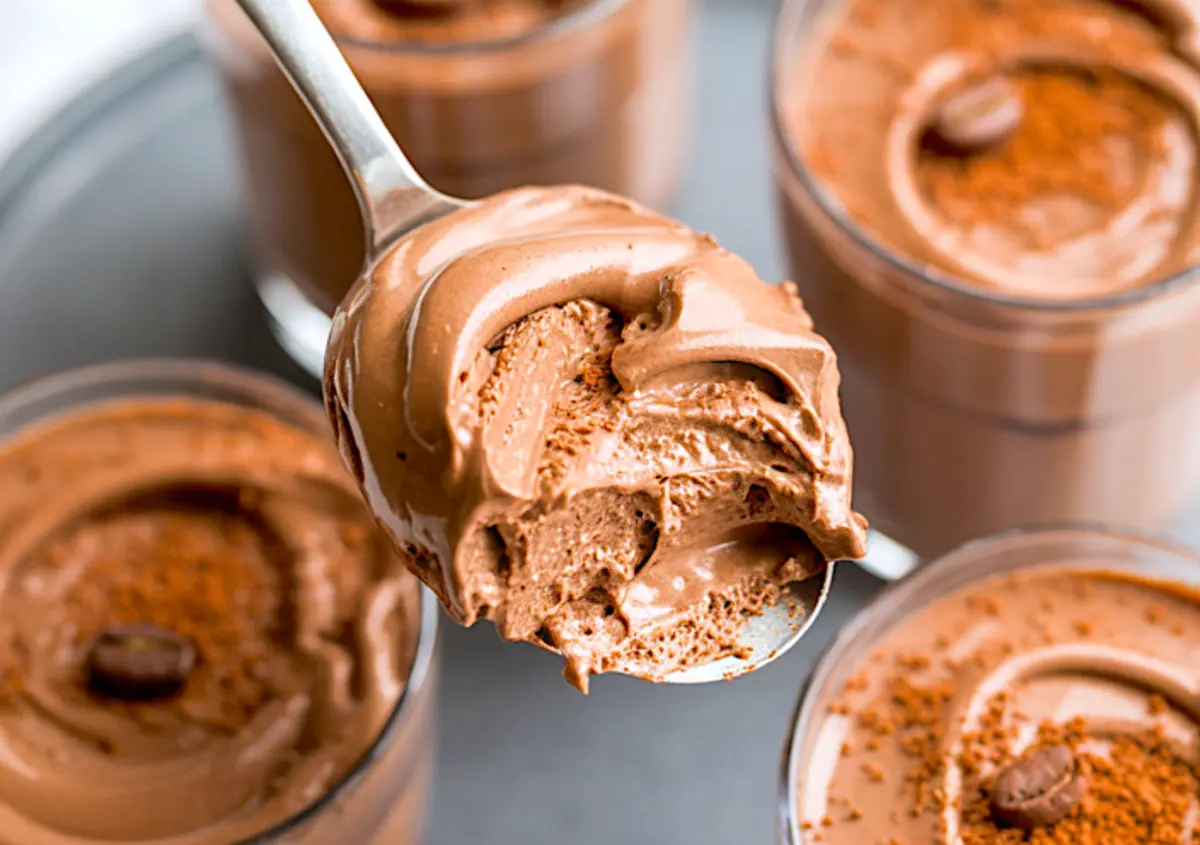 Spoonful of airy mocha mousse held above multiple glasses, showing creamy texture and chocolate coffee dessert layers with cocoa and coffee bean garnish.