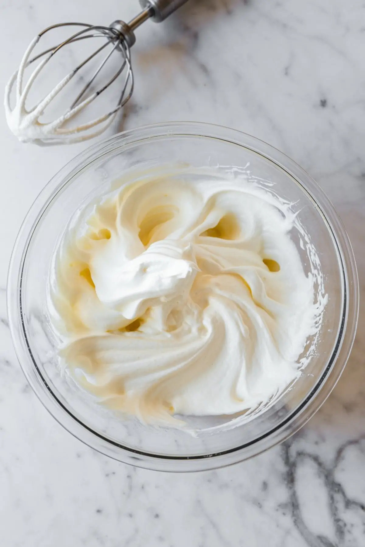 Fluffy whipped cream in a glass bowl on a marble countertop with a whisk attachment nearby, showing soft peaks texture ideal for desserts and mousse recipes.