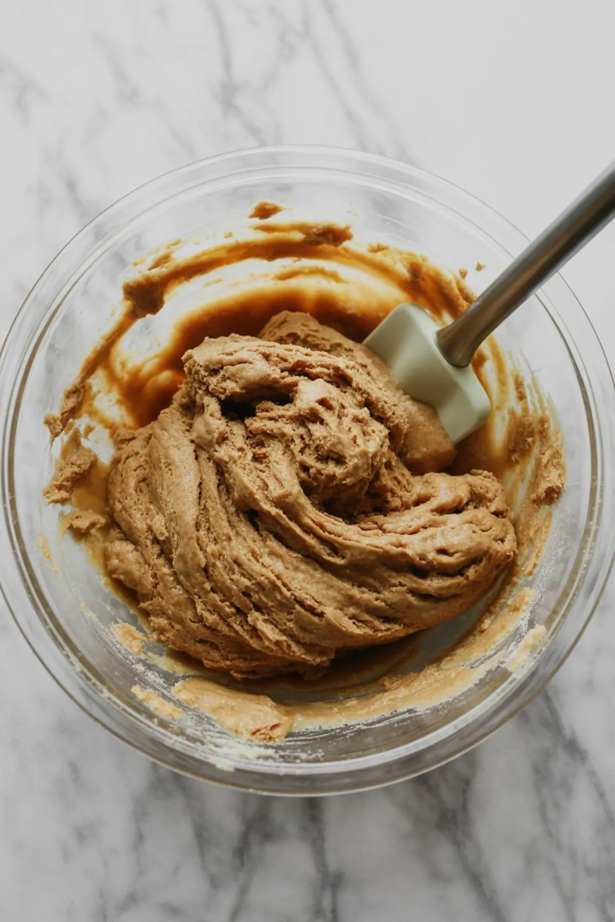 Thick ginger cookie dough being mixed with a silicone spatula in a glass bowl, set on a white marble backgroun