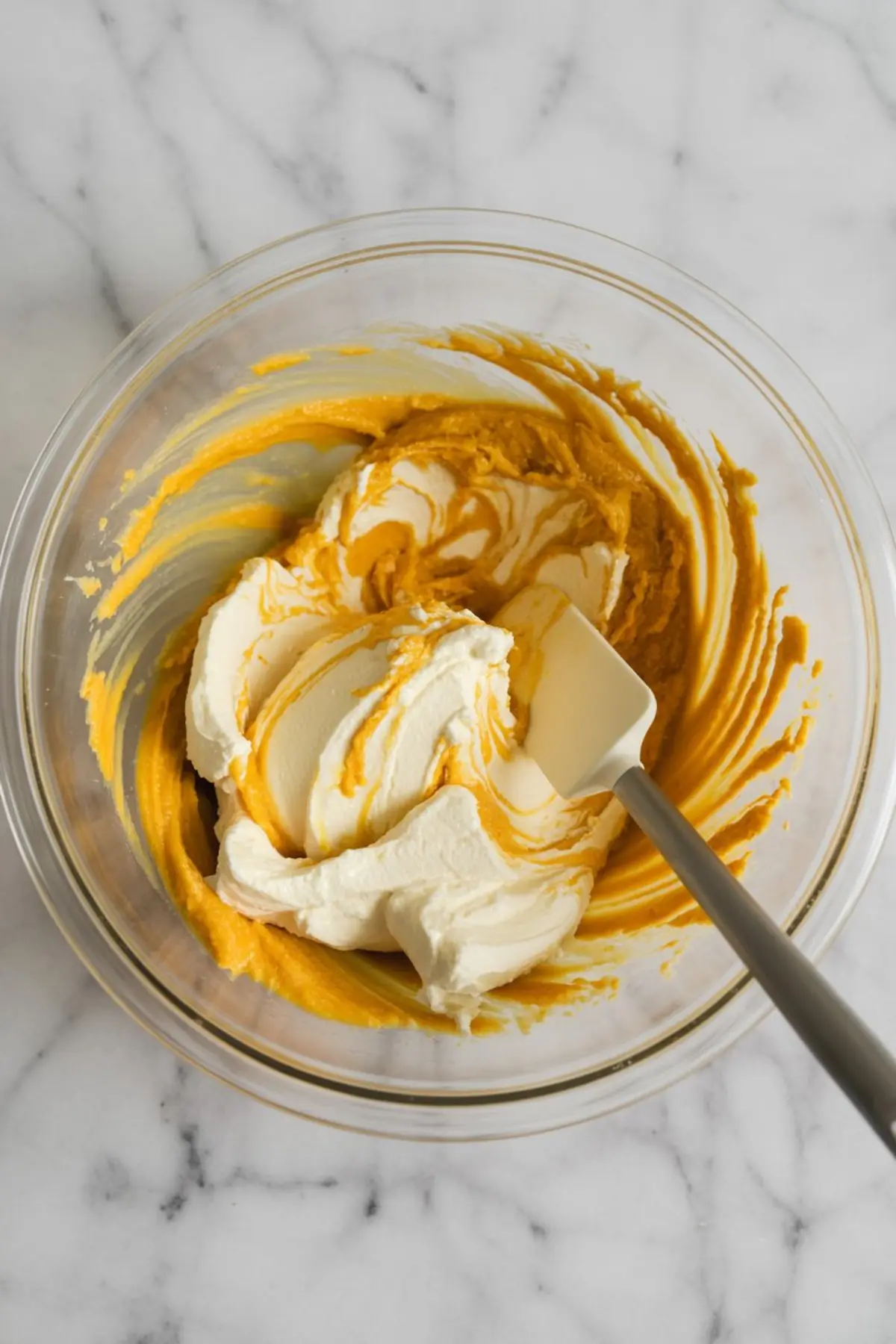 Mixing bowl with swirled pumpkin puree and cream cheese filling, being combined with a white spatula on a marble countertop.