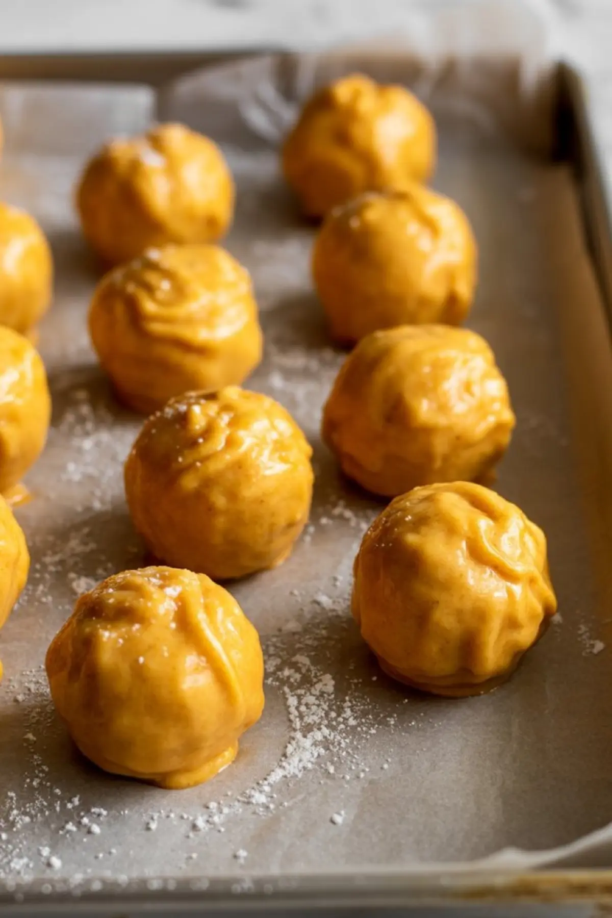Round pumpkin cheesecake truffles coated in orange-colored mixture arranged on a parchment-lined baking sheet with a light dusting of powdered sugar.