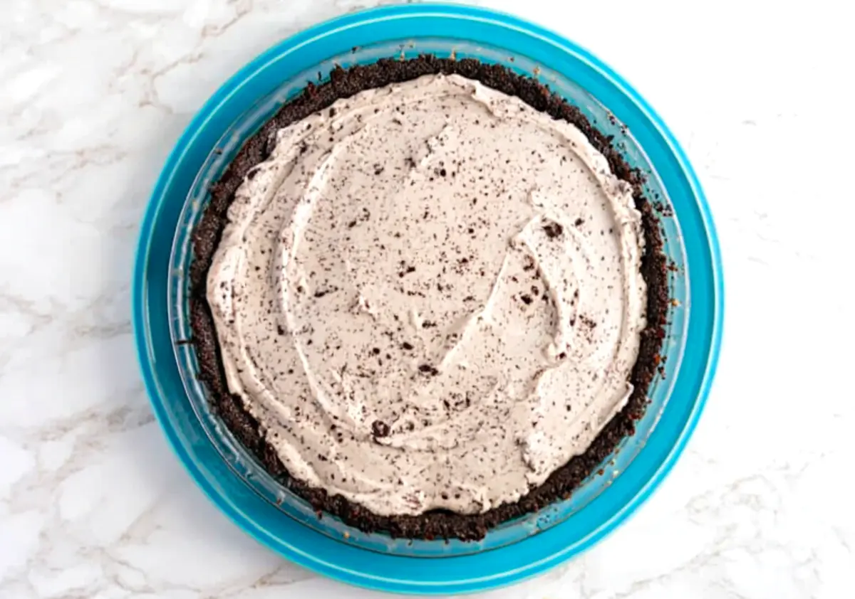 Overhead view of an Oreo cream pie in a chocolate cookie crust on a blue plate, topped with swirled cream filling and crushed cookie crumbs, ready to chill or serve.
