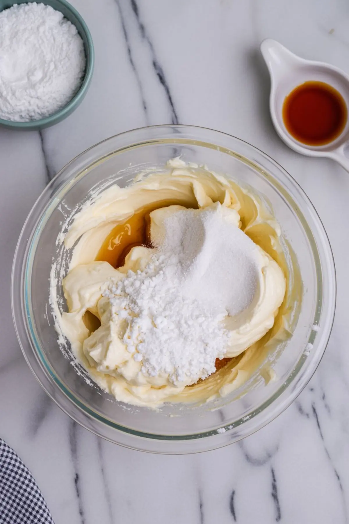 Glass bowl filled with cream cheese, powdered sugar, and vanilla extract on a marble surface, showing the beginning stages of no-bake Oreo pie filling preparation.