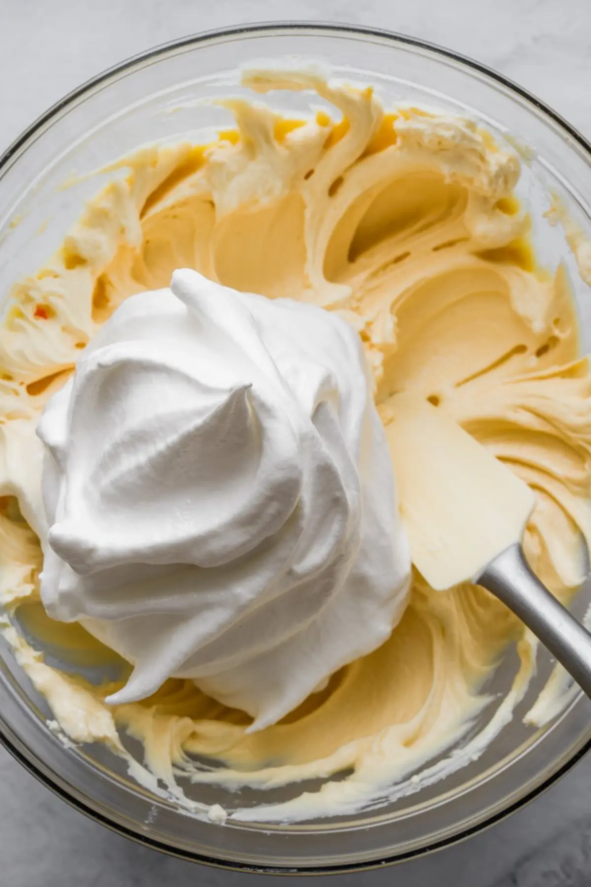 Mixing bowl containing a whipped cream topping being folded into a creamy mixture using a white silicone spatula, illustrating the light and fluffy pie filling process.