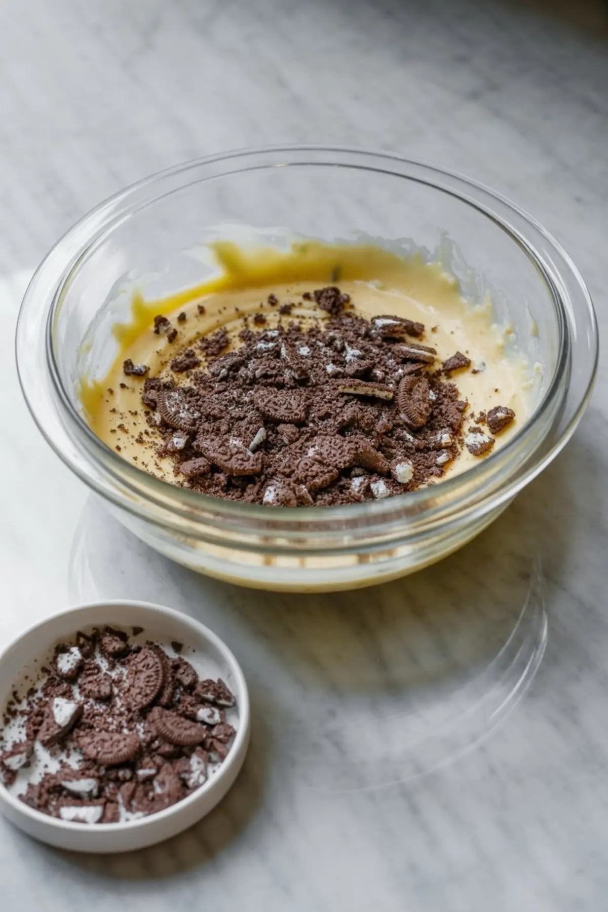 Crushed chocolate sandwich cookies being added to a yellow cream mixture in a glass bowl, capturing the step of incorporating cookies into the Oreo pie filling.