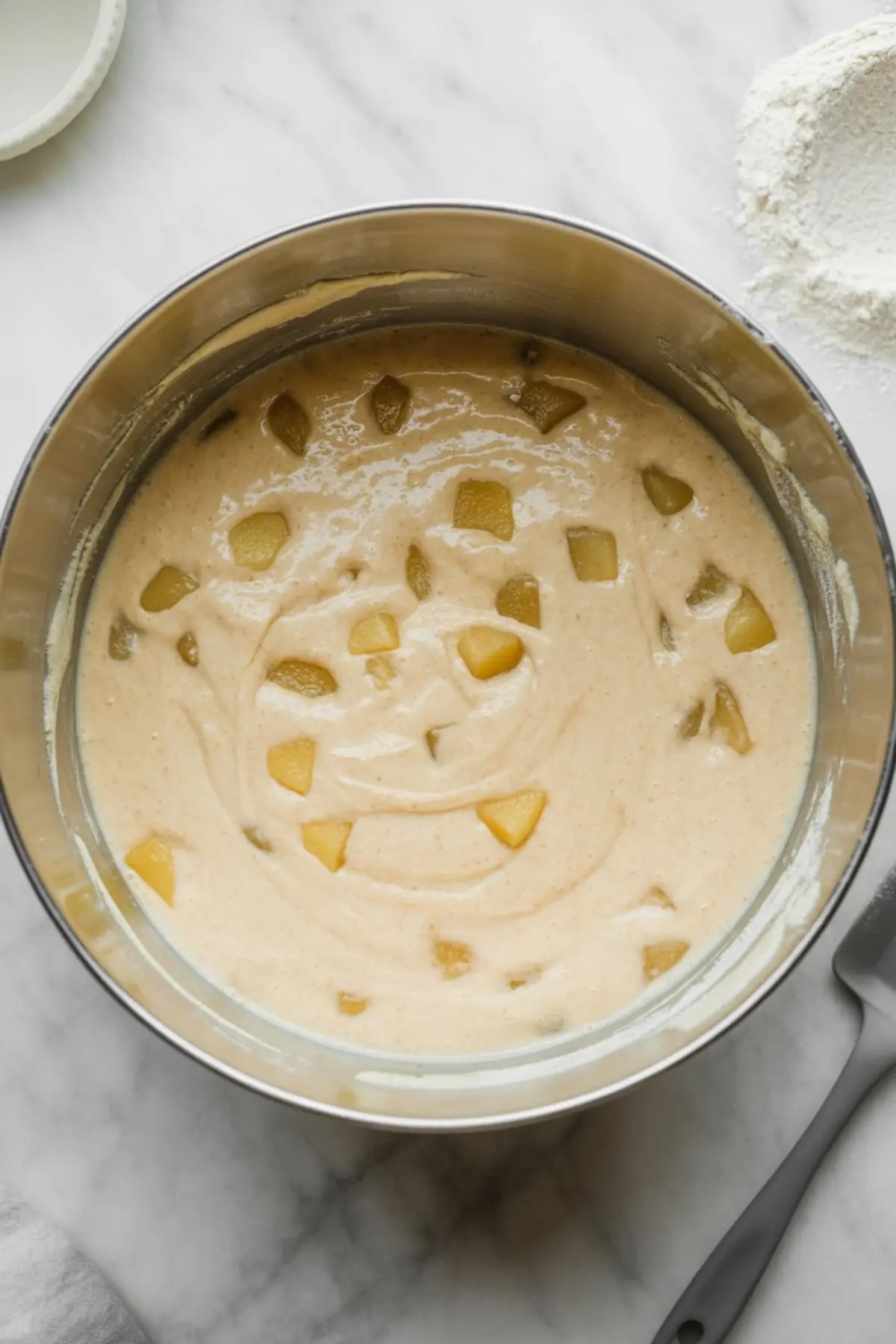 Mixing bowl containing pear cake batter with chunks of fresh pear, ready for baking, on a marble surface.