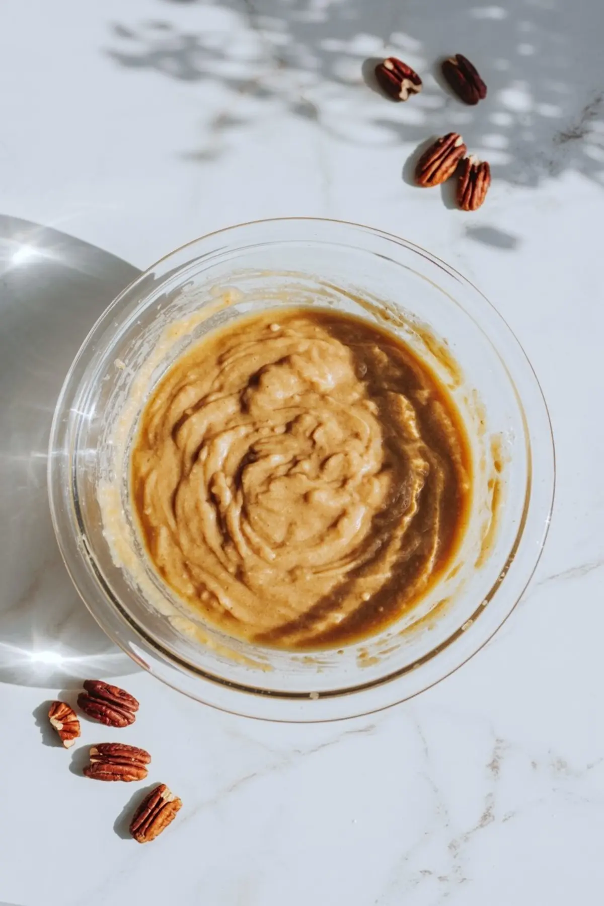 Glass bowl filled with mixed pecan pie filling batter placed on a white marble counter, surrounded by scattered whole pecans and natural shadows.