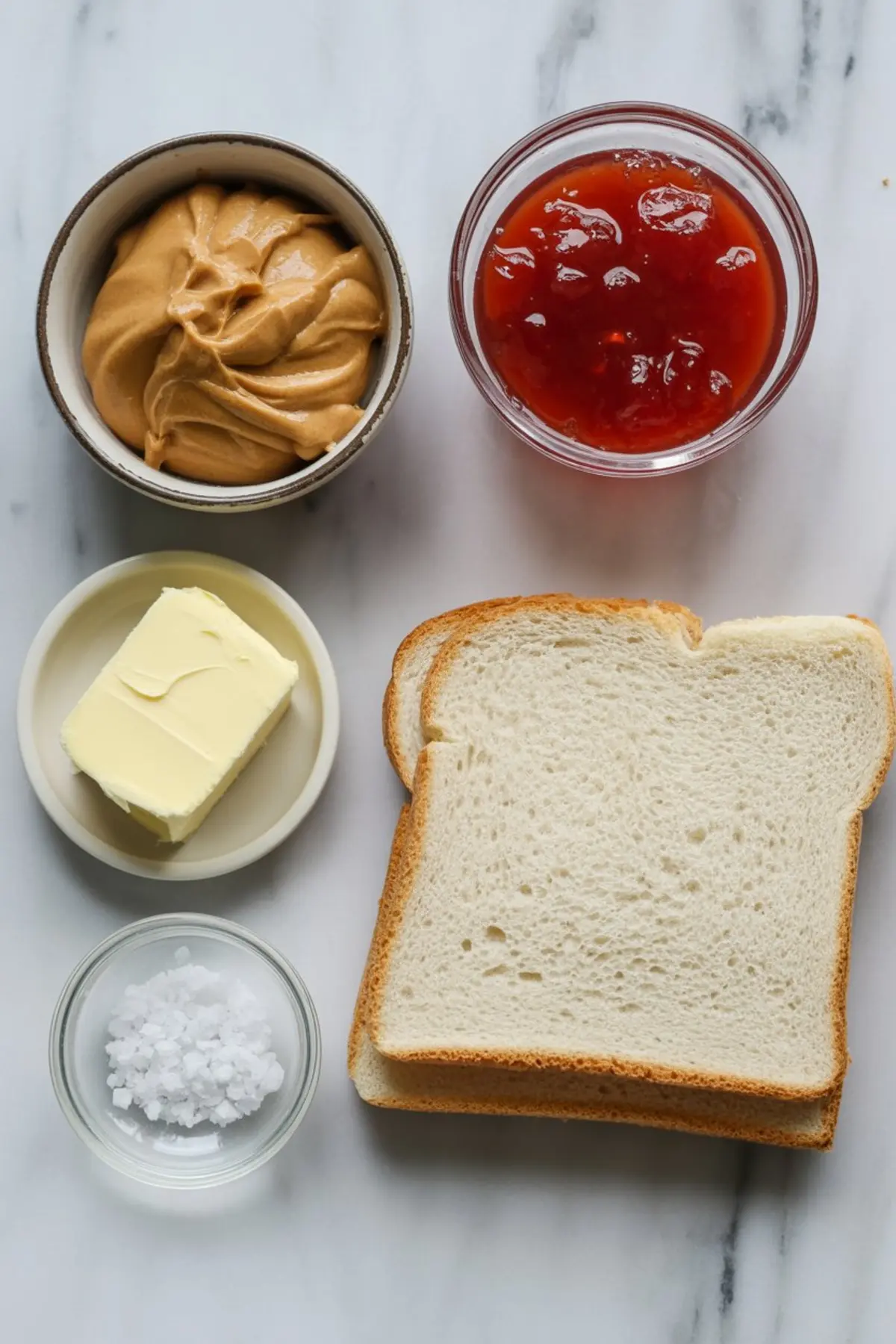 Top-down image of sandwich ingredients arranged neatly, including white bread, creamy peanut butter, strawberry jam, butter, and flaky sea salt on a white marble background.