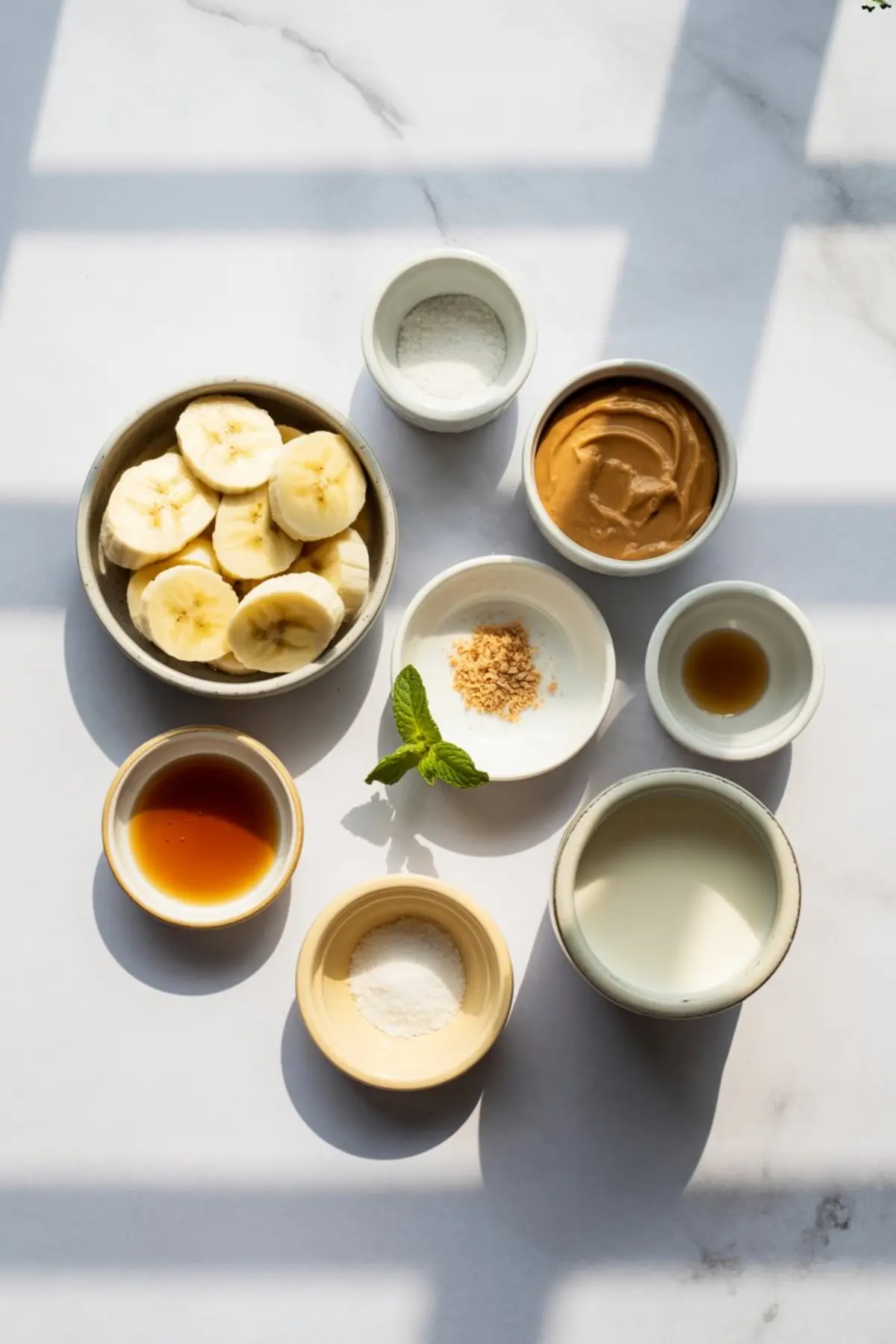 Flat lay of sliced bananas, creamy peanut butter, milk, vanilla, honey, and salt arranged in small ceramic bowls on sun-lit marble, ready for homemade peanut butter banana ice cream.