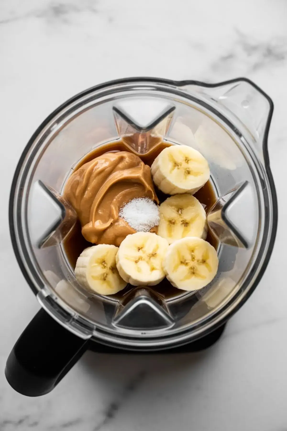 Overhead view of a blender jar filled with banana coins, peanut butter, maple syrup, and a pinch of salt, poised to blend into silky banana peanut butter nice cream.