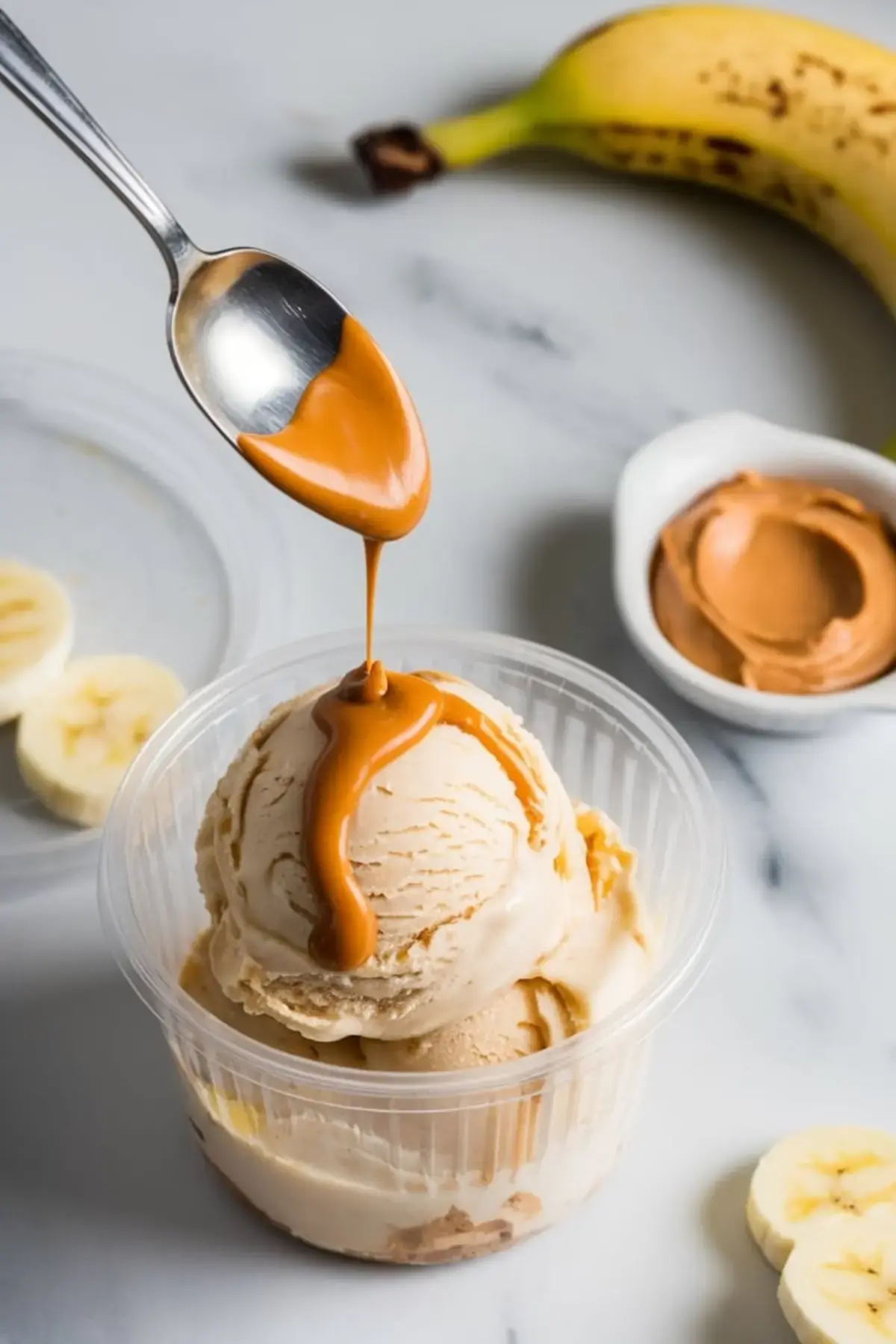 Metal spoon pours warm peanut butter over a single scoop of banana ice cream in a clear cup, with a ripe banana and peanut butter dish softly blurred in the background.