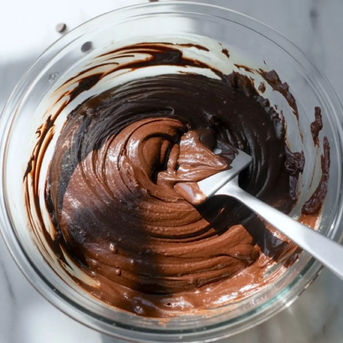 Thick chocolate brownie batter swirled in a glass bowl with a metal spatula, capturing a rich, glossy texture ready for baking.

