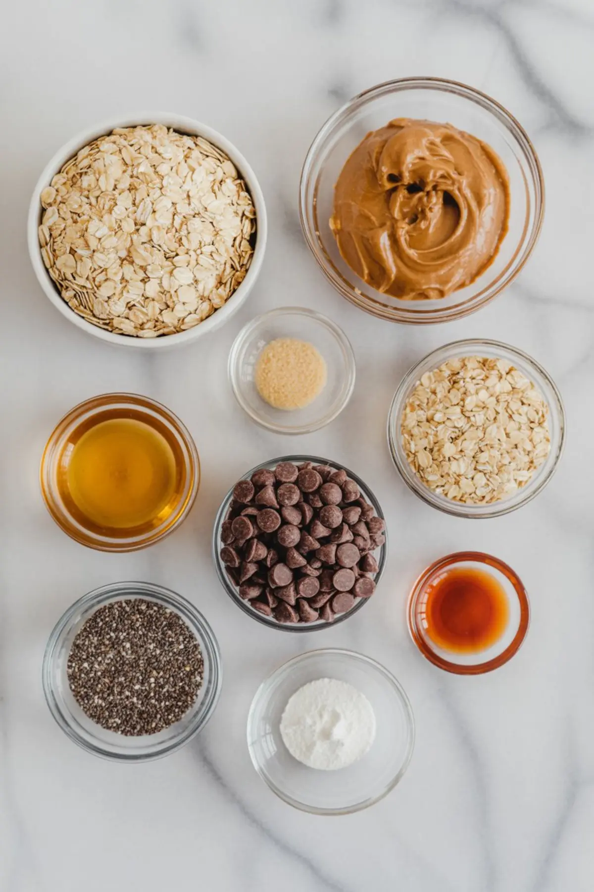 Flat lay of individual oat ball ingredients: peanut butter, rolled oats, chocolate chips, chia seeds, maple syrup, vanilla extract, protein powder, and brown sugar on white marble.