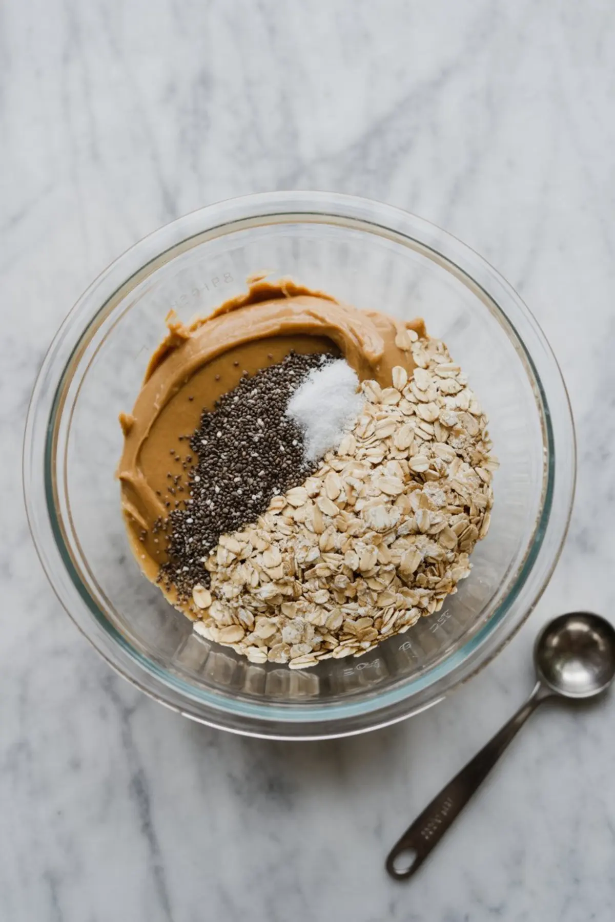 Glass bowl with creamy peanut butter, rolled oats, chia seeds, and a pinch of salt on a marble surface for a no-bake snack base.