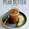 Close-up of a glass jar filled with smooth, homemade pear butter on a dark plate, garnished with fresh pear slices and a vintage spoon.